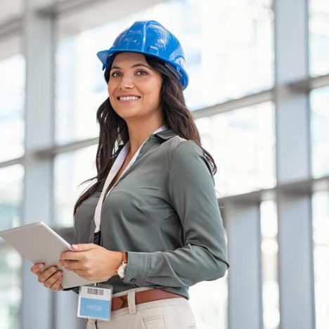 Beautiful mid adult woman architect wearing blue hardhat at construction site while working on digital tablet. Supervisor wearing safety helmet while working in a building site. Successful and proud inspector looking away with copy space.